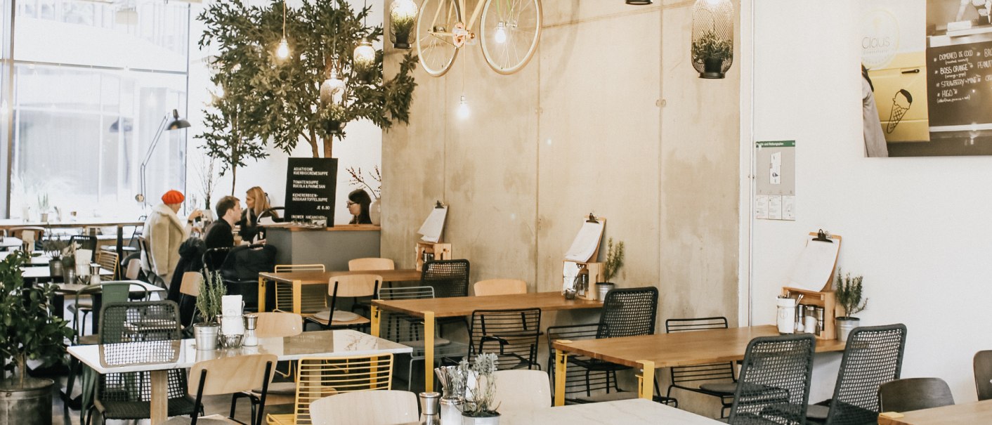 Modern café with a hanging bicycle on the wall, plants and cozy seating areas. People sit and chat at tables., © Claus Stuttgart