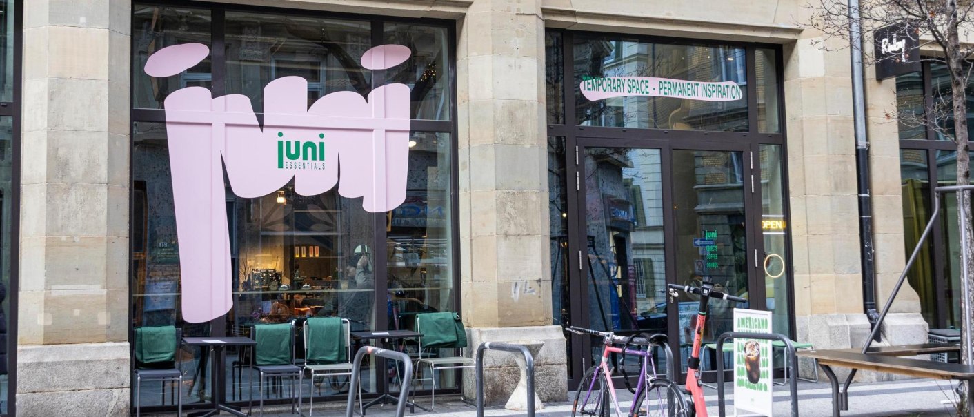 A café with large windows and the words 'juni essentials'. In front of it are bicycles and tables with chairs., © SMG, Sarah Schmid A café with large windows and the words 'juni essentials'. In front of it are bicycles and tables with chairs., © SMG, Sarah Schmid