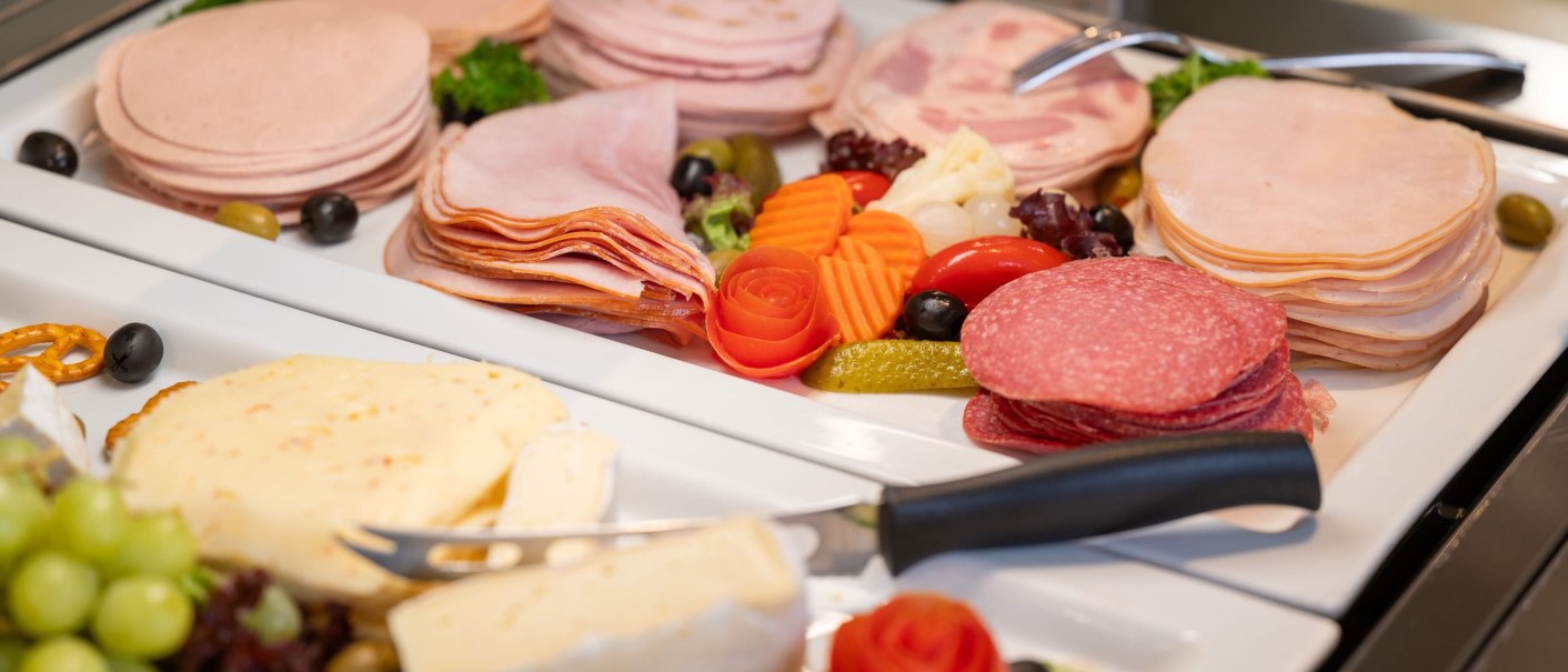 A breakfast buffet with a variety of cold meats and cheeses, decorated with vegetables and olives on white platters., &copy; Erikson Hotel