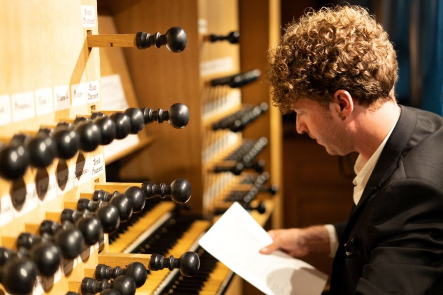 Ein Mann in einem Anzug spielt an einer Orgel und hält ein Blatt Papier in der Hand. Die Orgelregister sind deutlich sichtbar., © Johannes Schaugg