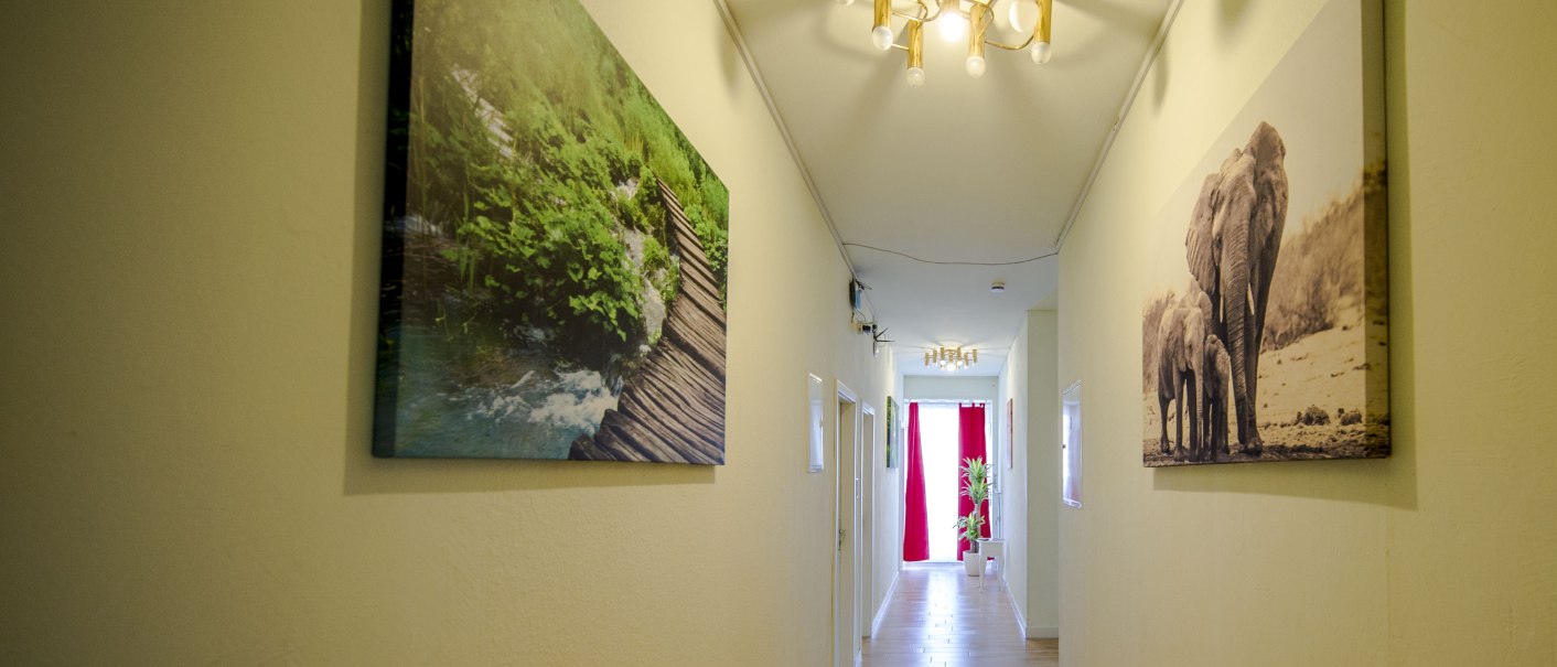 Ein heller Hotelkorridor mit Holzboden, dekoriert mit Natur- und Elefantenbildern. Eine moderne Deckenleuchte und rote Vorhänge am Ende des Flurs., © Hotel Ri