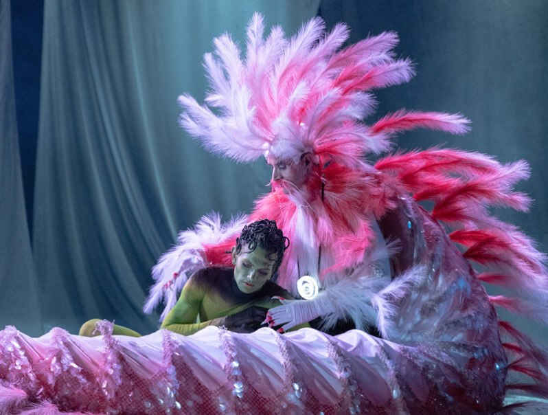 Zwei Personen in fantasievollen Kostümen auf einer Bühne. Eine Person trägt ein auffälliges Federkostüm in Rosa, die andere ein grünes Outfit., © Württembergische Staatstheater Stuttgart Zwei Personen in fantasievollen Kostümen auf einer Bühne. Eine Person trägt ein auffälliges Federkostüm in Rosa, die andere ein grünes Outfit., © Württembergische Staatstheater Stuttgart