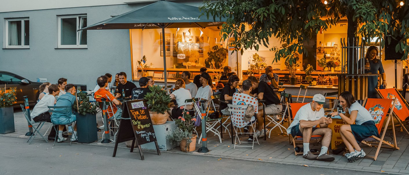 Abendliche Szene auf einer Terrasse mit vielen Menschen, die unter einem Sonnenschirm sitzen und sich unterhalten. Im Hintergrund leuchtet ein Café., © Radcafé Fietsen