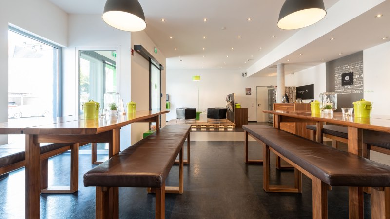 Modern lobby with wooden tables and benches, large lamps and a seating area in the background. Bright, inviting atmosphere., © a2 HOTELS Modern lobby with wooden tables and benches, large lamps and a seating area in the background. Bright, inviting atmosphere., © a2 HOTELS