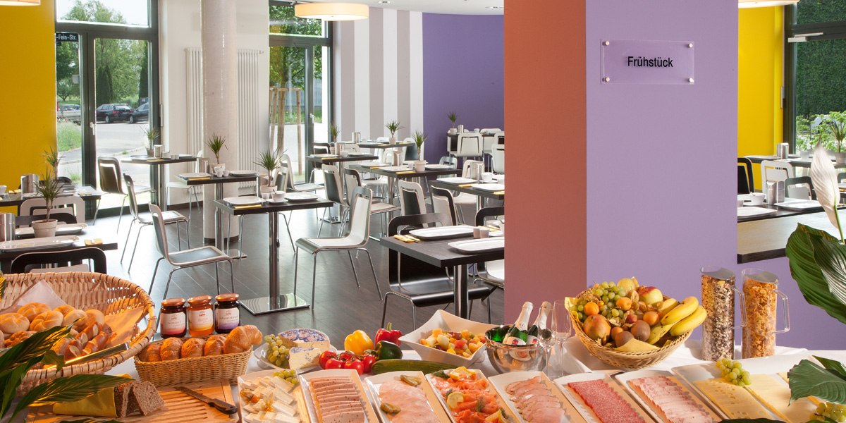 Ein reichhaltiges Frühstücksbuffet mit Brot, Aufschnitt, Obst und Müsli in einem modernen Restaurant mit bunten Wänden., © Star Inn Stuttgart Airport-Messe Ein reichhaltiges Frühstücksbuffet mit Brot, Aufschnitt, Obst und Müsli in einem modernen Restaurant mit bunten Wänden., © Star Inn Stuttgart Airport-Messe