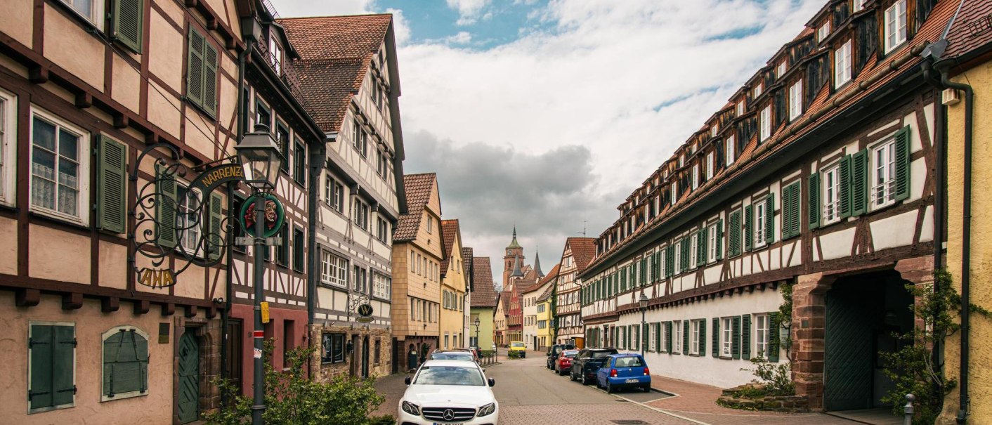 Half-timbered houses line a street in the old town of Weil der Stadt. Cars are parked along the street while clouds cover the sky., © Stuttgart-Marketing GmbH, Sarah Schmid
