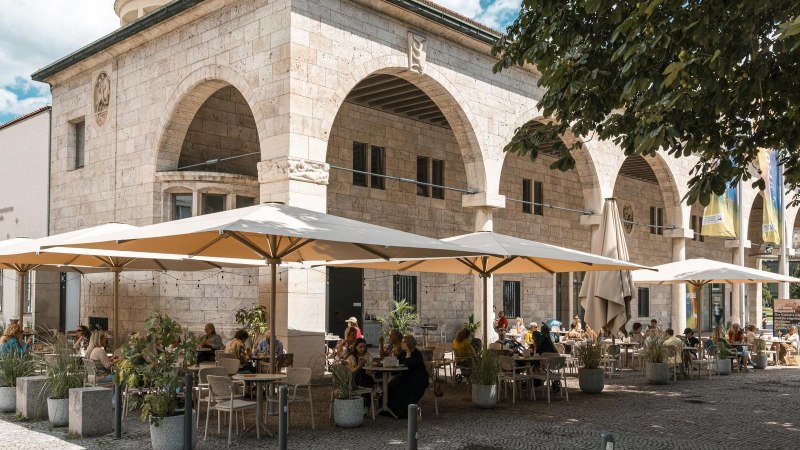 Ein Caf&eacute; mit gro&szlig;en Sonnenschirmen und G&auml;sten, die drau&szlig;en an Tischen sitzen. Das Geb&auml;ude hat B&ouml;gen und ist aus Stein., &copy; Stuttgart Marketing GmbH, Sarah Schmid