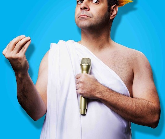 Mann in weißer Toga mit goldener Lorbeerkrone und Mikrofon, vor blauem Hintergrund. Er gestikuliert mit der Hand., © Theaterhaus Stuttgart e.V. Mann in weißer Toga mit goldener Lorbeerkrone und Mikrofon, vor blauem Hintergrund. Er gestikuliert mit der Hand., © Theaterhaus Stuttgart e.V.
