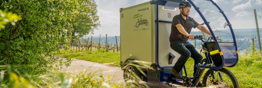 A man rides a cargo bike on a rural road, surrounded by green nature and trees. The sky is slightly cloudy., &copy; 70469R!
