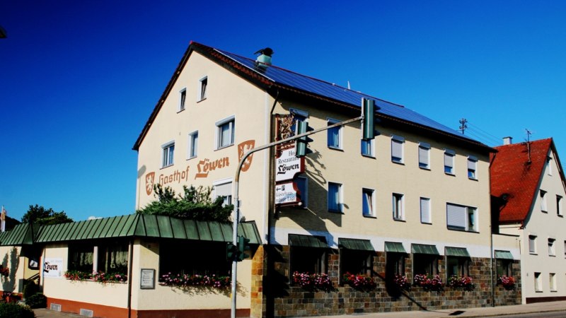 Der Gasthof Löwen mit beiger Fassade und roten Dächern, dekoriert mit Blumen, unter blauem Himmel., © Ulli Kellenbenz Der Gasthof Löwen mit beiger Fassade und roten Dächern, dekoriert mit Blumen, unter blauem Himmel., © Ulli Kellenbenz