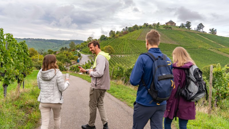 Menschen wandern auf einem Weinweg, halten Weingläser. Im Hintergrund Weinberge und die Grabkapelle auf einem Hügel., © Stuttgart-Marketing GmbH, Martina Denker