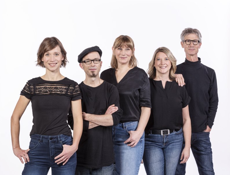 F&uuml;nf Personen in schwarzer Kleidung stehen nebeneinander und l&auml;cheln in die Kamera. Der Hintergrund ist wei&szlig;., &copy; Improtheater Stuttgart / Rainer Wengel Fotografie