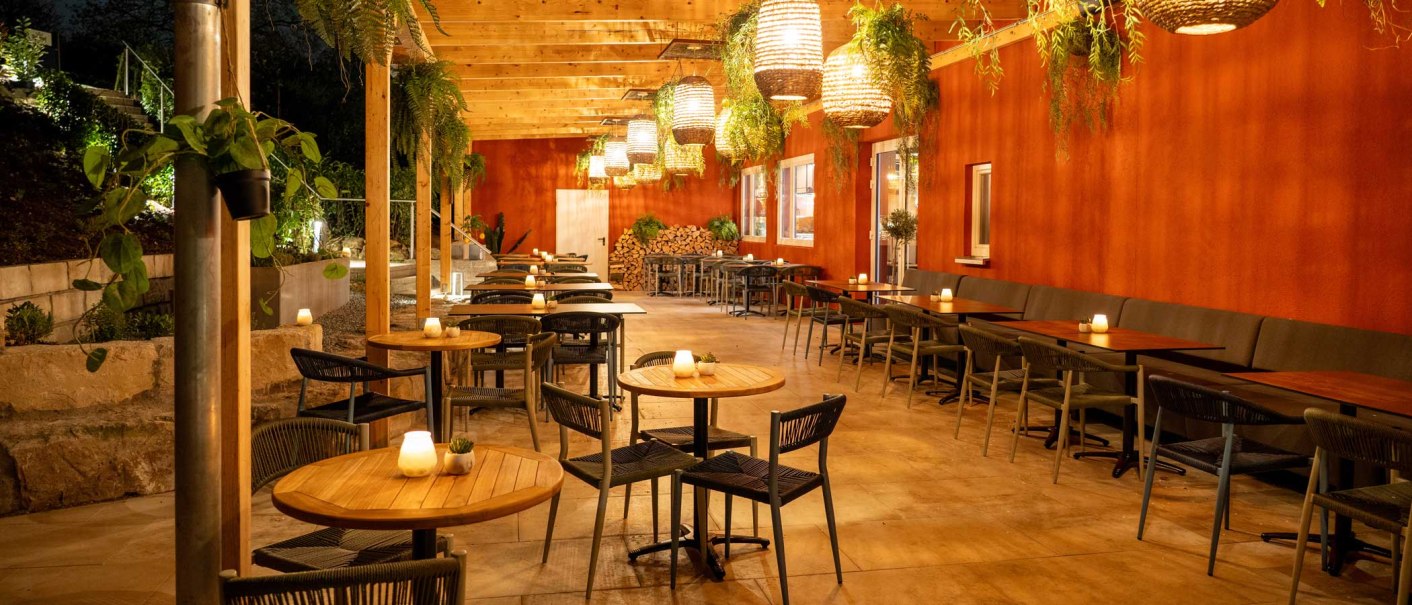 Gemütlicher Außenbereich mit Holztischen, Stühlen und hängenden Pflanzen. Warme Beleuchtung und rote Wand schaffen eine einladende Atmosphäre., © SatzDrei