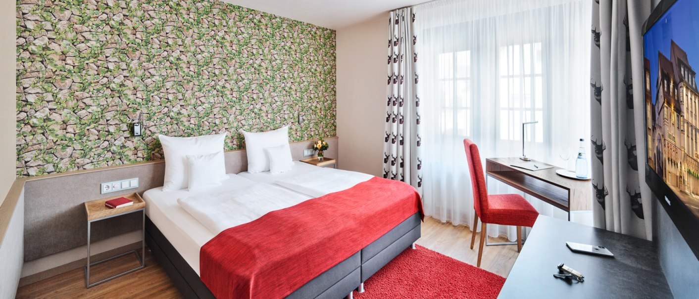 Modernes Hotelzimmer mit Doppelbett, rotem Teppich, Schreibtisch und Fenster mit Vorhängen. Die Wand hat eine grüne Steinoptik-Tapete., © Hirsch Hotel