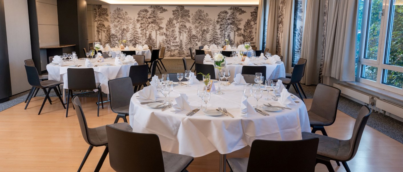 Elegant breakfast room with round, white-clothed tables, black chairs and a wall with forest wallpaper. Large windows let in natural light., © Erikson Hotel