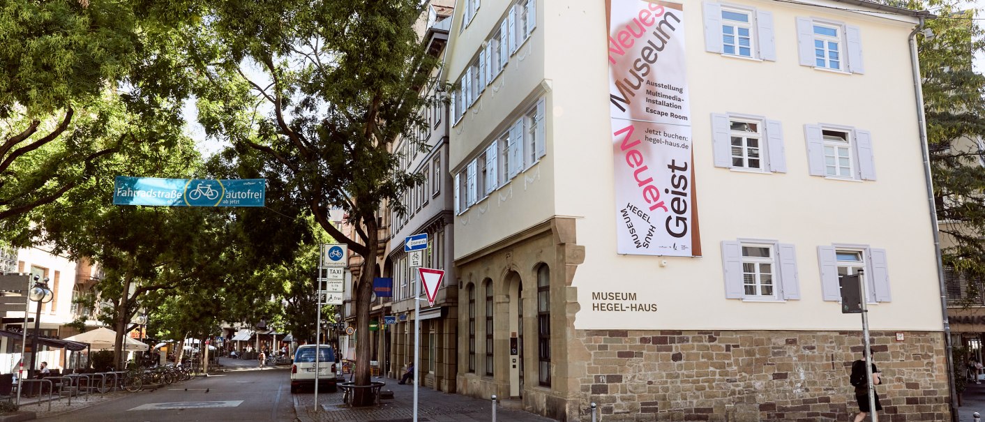 Das Hegel-Haus in Stuttgart mit einem großen Banner an der Fassade. Die Straße davor ist autofrei und von Bäumen gesäumt., © Julia Ochs Das Hegel-Haus in Stuttgart mit einem großen Banner an der Fassade. Die Straße davor ist autofrei und von Bäumen gesäumt., © Julia Ochs