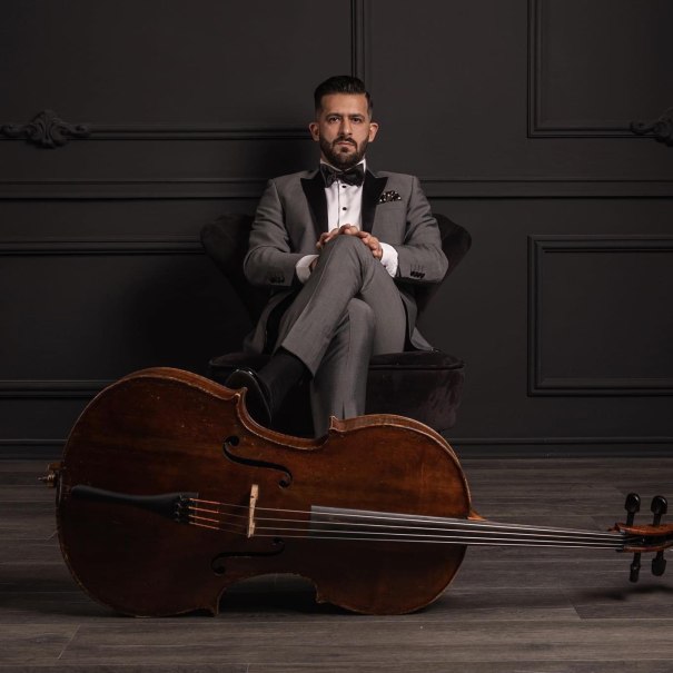 Ein elegant gekleideter Mann im grauen Anzug sitzt vor einer dunklen Wand. Ein Cello liegt vor ihm auf dem Boden., &copy; Elem&eacute;r Duka