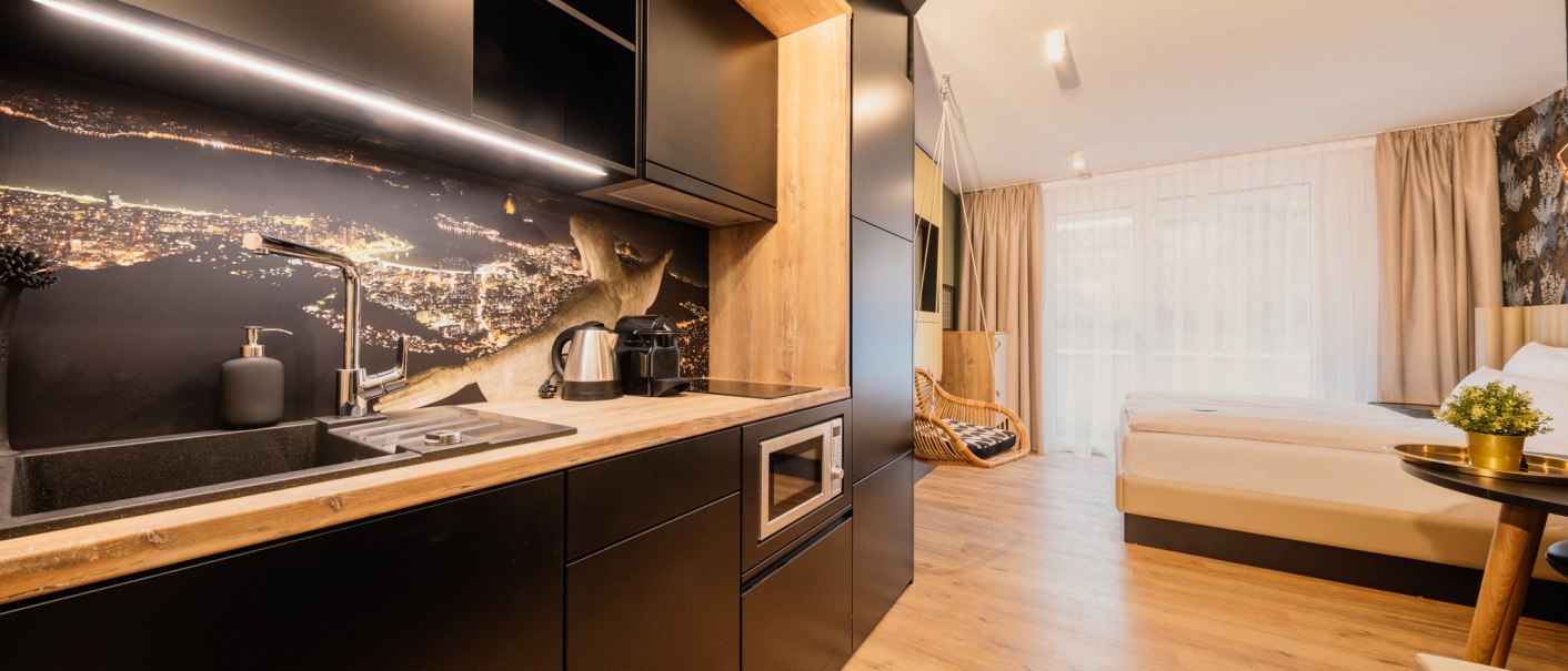 Modern apartment with open kitchen, wooden floor and cozy sleeping area. Black kitchen cabinets and a large bed dominate the room., © Rioca Stuttgart Posto 4 GmbH Modern apartment with open kitchen, wooden floor and cozy sleeping area. Black kitchen cabinets and a large bed dominate the room., © Rioca Stuttgart Posto 4 GmbH