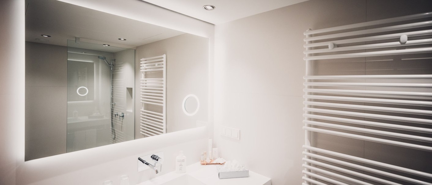Modernes Badezimmer mit großem Spiegel, Waschbecken, Handtuchheizkörper und Duschbereich. Helle Beleuchtung und minimalistische Einrichtung., © Abacco Hotels GmbH Modernes Badezimmer mit großem Spiegel, Waschbecken, Handtuchheizkörper und Duschbereich. Helle Beleuchtung und minimalistische Einrichtung., © Abacco Hotels GmbH