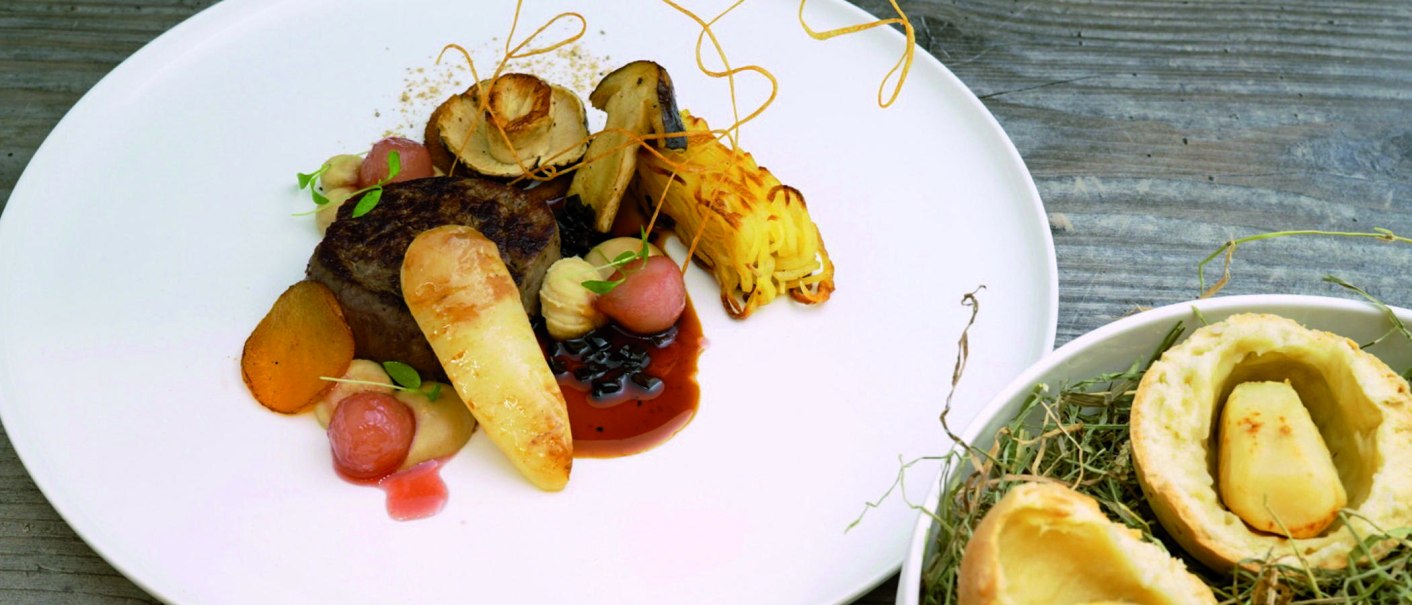 Gourmetgericht auf weißem Teller: Fleisch, Gemüse, Soße und kunstvolle Garnitur. Daneben ein Schälchen mit Gebäck und Kartoffel auf Holzuntergrund., © Speisemeisterei