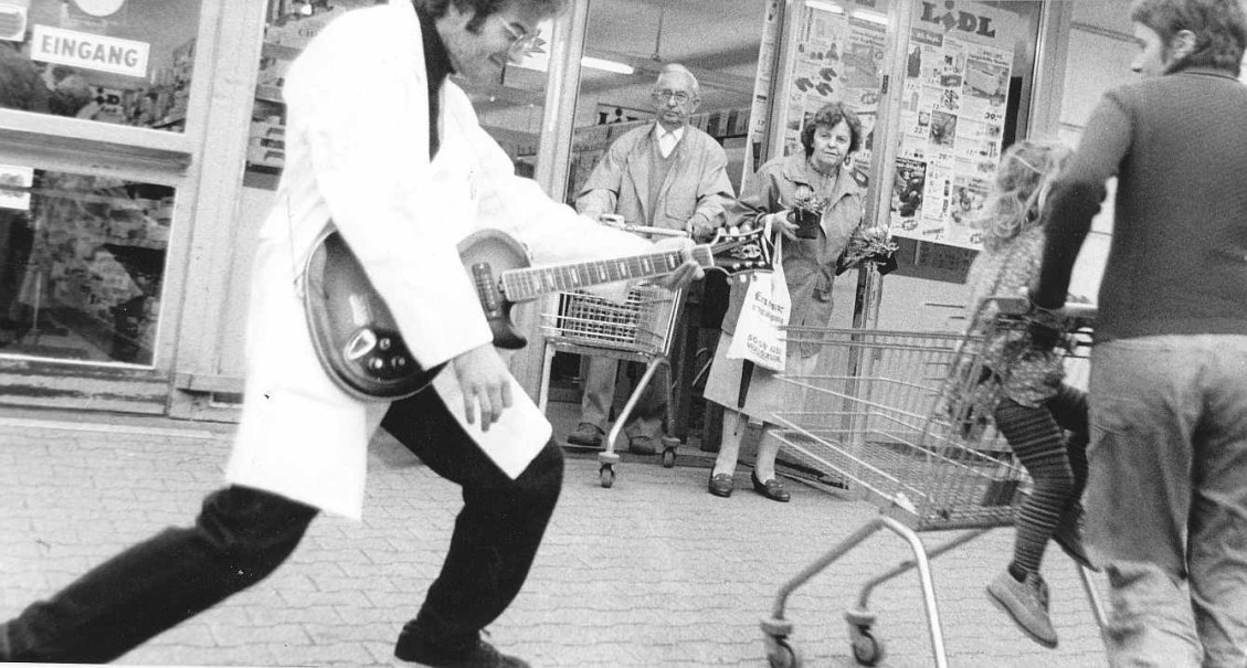 Ein Mann in einem wei&szlig;en Mantel spielt Gitarre vor einem Supermarkt. Passanten mit Einkaufswagen beobachten die Szene interessiert., &copy; Rock&rsquo;n&rsquo;Roll Diktator