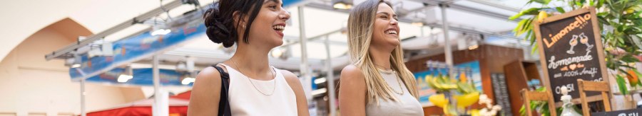 Two women laugh in the Stuttgart market hall, surrounded by market stalls with fresh produce and a limoncello sign., &copy; &copy; Stuttgart-Marketing GmbH, Ingolf Pompe