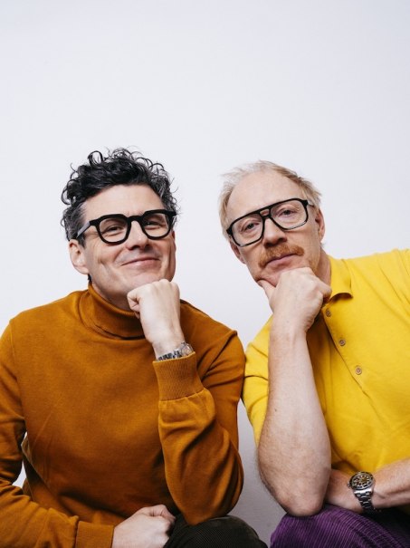 Two men with glasses and colorful sweaters pose smiling in front of a white wall. Both are resting their chins on their hands., © Theaterhaus Stuttgart e.V. Two men with glasses and colorful sweaters pose smiling in front of a white wall. Both are resting their chins on their hands., © Theaterhaus Stuttgart e.V.
