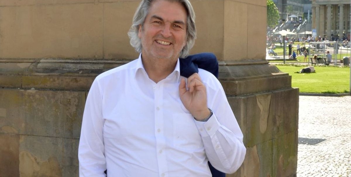 Ein Mann in weißem Hemd steht lächelnd vor einem historischen Gebäude auf einem sonnigen Platz., © Wolfgang Seljé