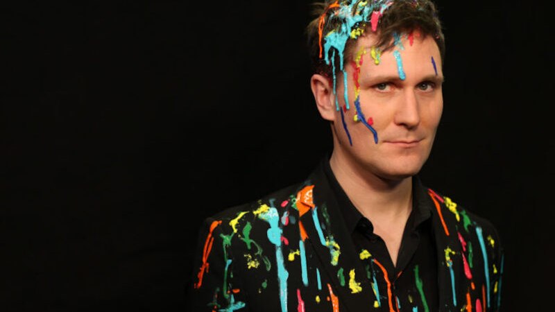 Eine Person mit bunten Farbspritzern auf Gesicht und Kleidung vor einem schwarzen Hintergrund., &copy; Rosenau Kultur e.V.