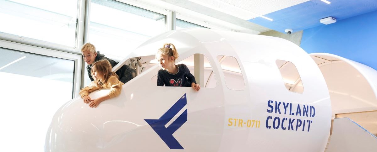 Drei Kinder spielen in einem Cockpit-Nachbau mit der Aufschrift 'SkyLand Cockpit' und blicken aus den Fenstern. Helle, moderne Umgebung., &copy; Flughafen Stuttgart