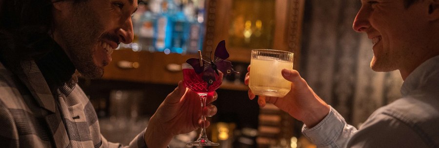Zwei M&auml;nner sto&szlig;en in einer Bar mit Cocktails an, einer in einem karierten Sakko, der andere in einem hellen Hemd. Im Hintergrund sind Flaschen zu sehen., &copy; SMG, Martina Denker