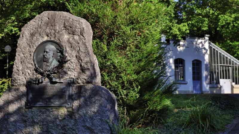 Gedenkstein f&uuml;r Gottlieb Daimler in einem gr&uuml;nen Park, rechts ein wei&szlig;es Geb&auml;ude mit Glasfenstern., &copy; Stuttgart-Marketing GmbH Achim Mende