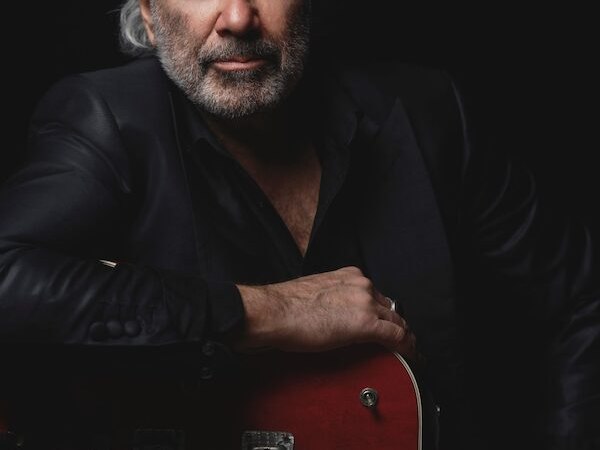 Ein Mann mit grauem Haar und Bart in schwarzem Anzug lehnt sich auf eine rote E-Gitarre vor schwarzem Hintergrund., © Theaterhaus Stuttgart e.V. Ein Mann mit grauem Haar und Bart in schwarzem Anzug lehnt sich auf eine rote E-Gitarre vor schwarzem Hintergrund., © Theaterhaus Stuttgart e.V.