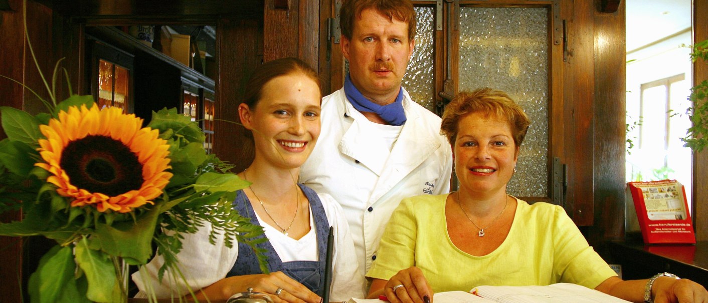 Drei Personen stehen an einer Rezeption: eine Frau in Tracht, ein Mann in Kochkleidung und eine Frau mit einem Buch. Eine Sonnenblume schmückt den Tresen., © R.Storr Drei Personen stehen an einer Rezeption: eine Frau in Tracht, ein Mann in Kochkleidung und eine Frau mit einem Buch. Eine Sonnenblume schmückt den Tresen., © R.Storr