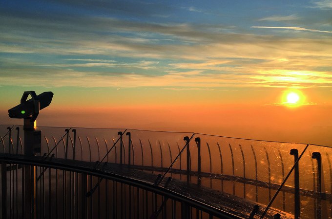 Aussichtsplattform des SWR Fernsehturms bei Sonnenaufgang. Ein Fernrohr steht neben einem Glasgeländer, während die Sonne am Horizont aufgeht., © SWR Media Services GmbH / SWR Fernsehturm Stuttgart