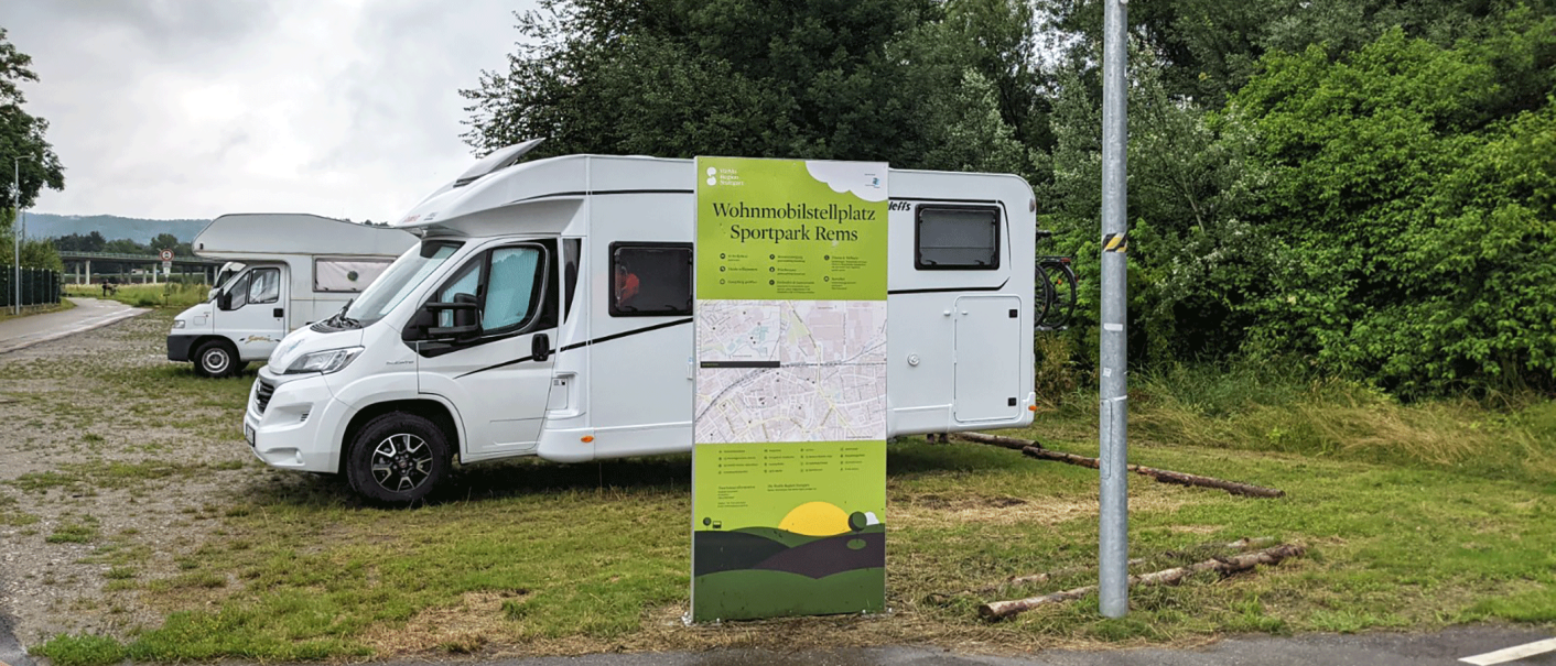 Zwei Wohnmobile parken auf einem Stellplatz im Sportpark Rems. Ein großes Schild informiert über den Standort. Im Hintergrund sind Bäume zu sehen., © Stadtverwaltung Schorndorf Zwei Wohnmobile parken auf einem Stellplatz im Sportpark Rems. Ein großes Schild informiert über den Standort. Im Hintergrund sind Bäume zu sehen., © Stadtverwaltung Schorndorf
