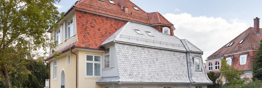 Zweistöckiges Haus mit rotem Ziegeldach, gelber Fassade und weißen Fenstern, umgeben von Bäumen und Rasen., © Philip Kottlorz