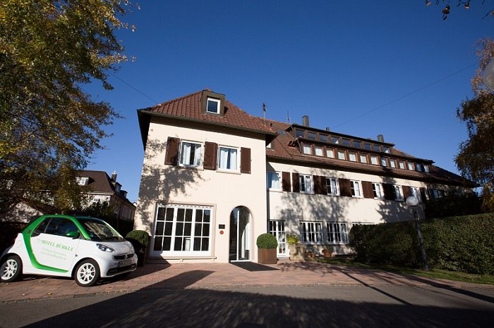 A two-story hotel with brown shutters, surrounded by trees. A small car is parked in front of it. The sky is clear and blue., &copy; Hotel B&uuml;rkle