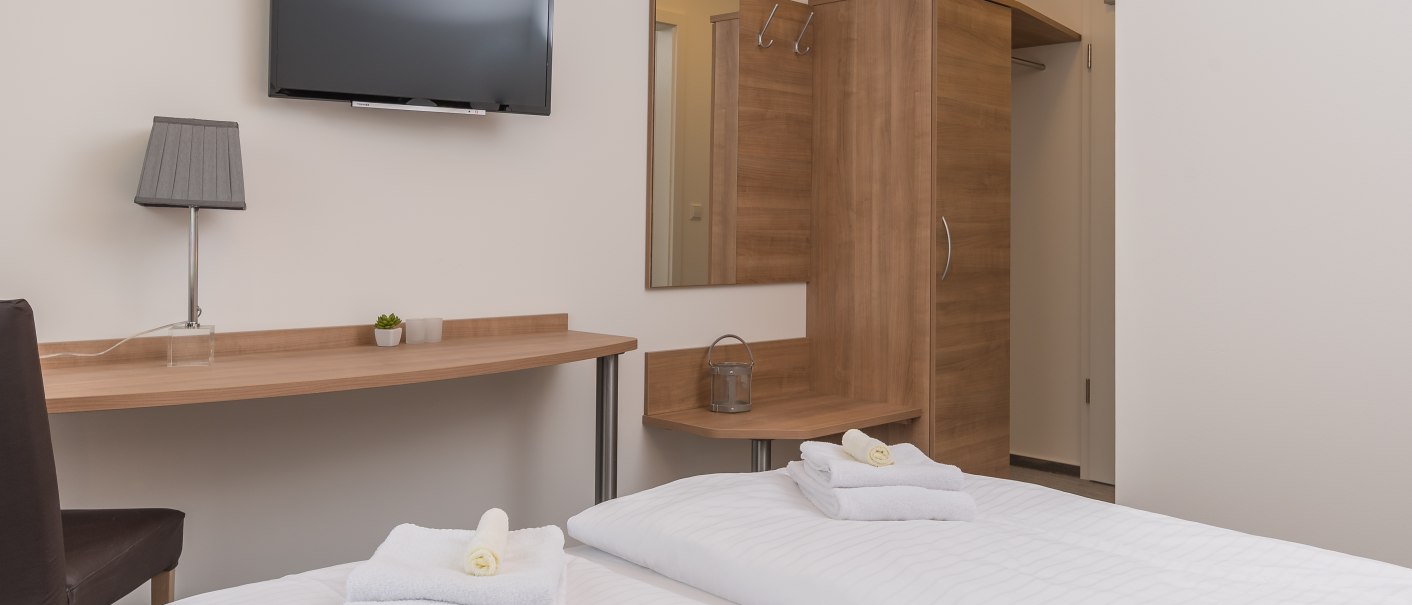 Hotelzimmer mit zwei Einzelbetten, Schreibtisch, Flachbildfernseher, Kleiderschrank und Lampe. Handtücher auf den Betten, minimalistische Einrichtung., © TOMAS