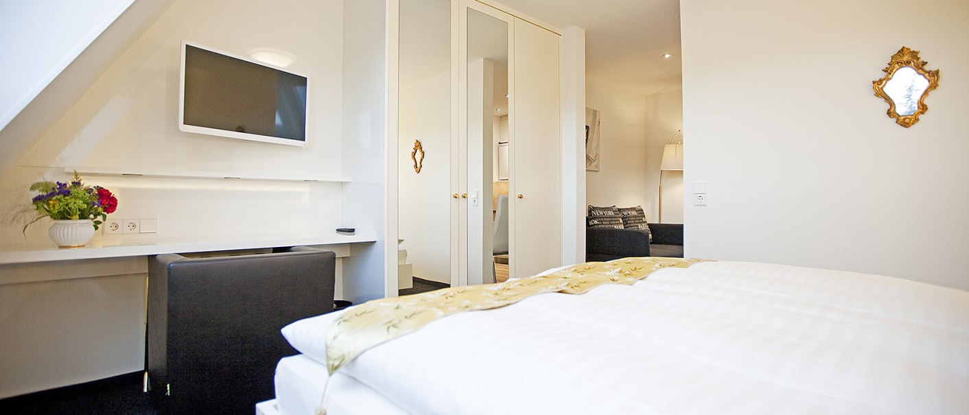 Helles Schlafzimmer mit Doppelbett, Flachbildfernseher, Spiegel und Blumen auf einem Tisch. Ein Schrank mit Spiegeln und ein Sofa im Hintergrund., &copy; Hotel Krone