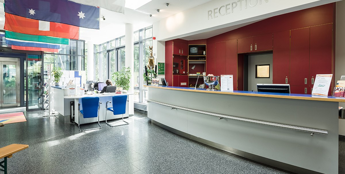 Bright entrance area with reception, red cupboards, blue chairs and flags on the ceiling., © TOMAS Bright entrance area with reception, red cupboards, blue chairs and flags on the ceiling., © TOMAS
