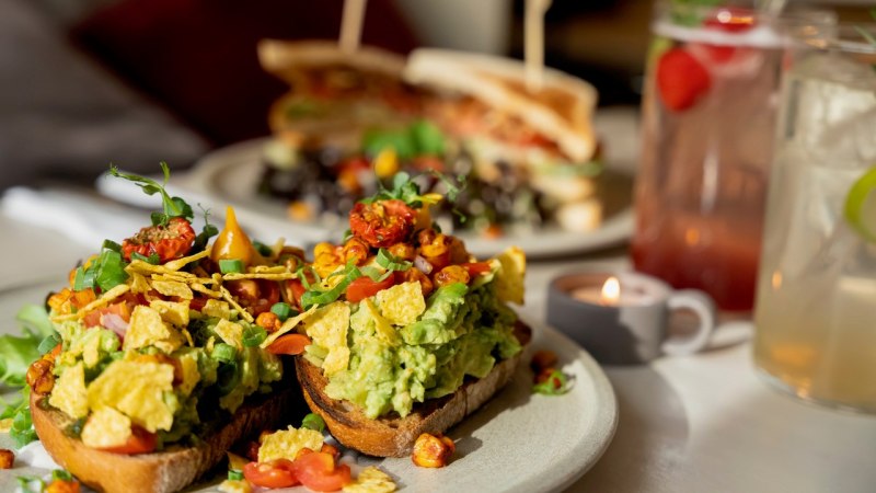 Avocado-Toast mit Tomaten und Kr&auml;utern, im Hintergrund ein Sandwich und Getr&auml;nke. Ein Teelicht sorgt f&uuml;r gem&uuml;tliche Atmosph&auml;re., &copy; Veganes Gold&reg;