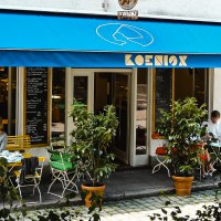 Ein Café mit blauer Markise und dem Namen 'Koenig'. Gäste sitzen draußen an Tischen und genießen ihre Getränke. Pflanzen dekorieren den Eingangsbereich., © KoenigX