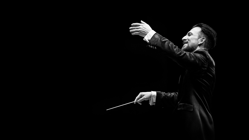 Ein Dirigent in einem schwarzen Anzug dirigiert mit einem Taktstock vor einem schwarzen Hintergrund., © Württembergische Staatstheater Stuttgart Ein Dirigent in einem schwarzen Anzug dirigiert mit einem Taktstock vor einem schwarzen Hintergrund., © Württembergische Staatstheater Stuttgart