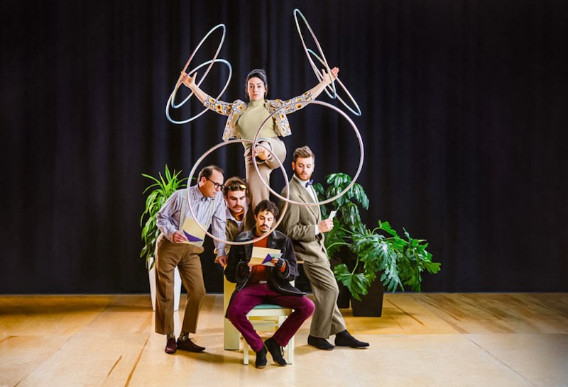 Eine Frau jongliert mit Reifen auf einem Stuhl, umgeben von vier M&auml;nnern, die konzentriert lesen. Pflanzen und ein dunkler Vorhang im Hintergrund., &copy; Theaterhaus Stuttgart e.V.