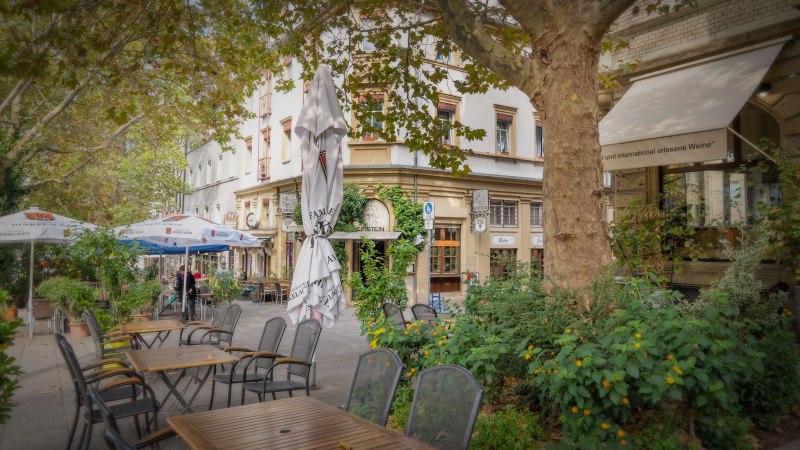 Ein Bistro am Wilhelmsplatz mit leeren Tischen und St&uuml;hlen im Freien, umgeben von gr&uuml;nen Pflanzen und B&auml;umen., &copy; Martina Denker