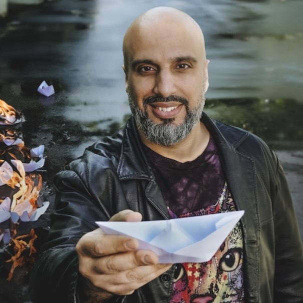 Ein Mann mit Glatze und Bart hält ein Papierboot. Im Hintergrund brennen weitere Boote auf einer nassen Straße., © Renitenztheater Stuttgart e.V.