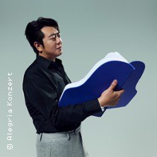 Ein Mann in schwarzem Hemd und grauer Hose hält ein großes, blaues Buch in Form eines Flügels., © links im Bild