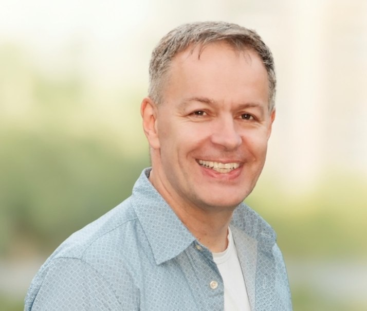 A man with gray hair and a blue shirt smiles at the camera. The background is blurred and greenish., &copy; Renitenztheater Stuttgart e.V.