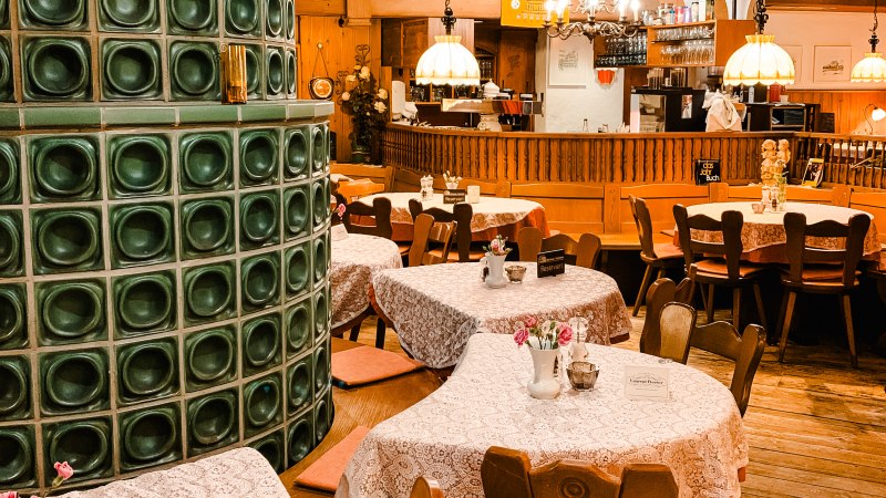 Cozy restaurant with wooden furniture, tablecloths and a green tiled stove. Lamps hanging from the ceiling, bar in the background., © Weinstube Kachelofen